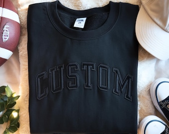 May include: Black sweatshirt with the word "CUSTOM" embroidered across the chest in a collegiate font. The sweatshirt is folded on a white, fluffy surface. A football, a baseball cap, and sneakers are also in the image.