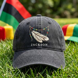 May include: A charcoal grey baseball cap featuring an embroidered design of two light-coloured feathers draped with a string of small red, yellow, and green beads. The word JACKSON is embroidered in white capital letters below the feathers.