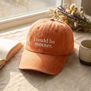 May include: Orange baseball cap with the embroidered text "i could be meaner." The cap has a curved brim and a button on top. The hat is sitting on a neutral-coloured surface.