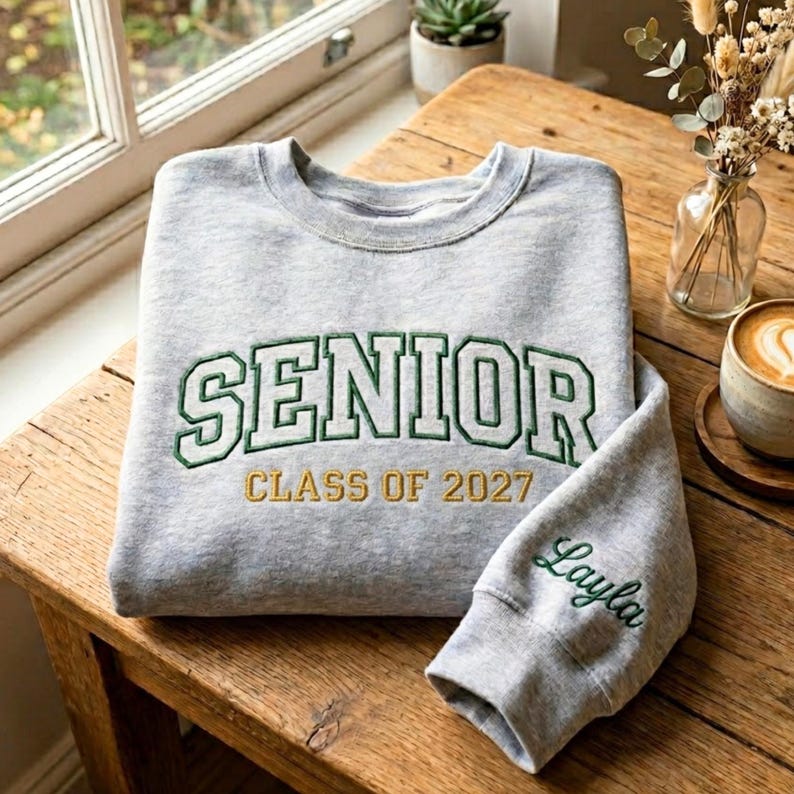 May include: A gray sweatshirt with the word "SENIOR" in green and white lettering, with "CLASS OF 2027" in gold lettering. The sleeve has the name "Layla" embroidered in green script.