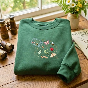 Könnte beinhalten: Ein waldgrüner Rundhals-Pullover mit einer Stickerei, die ein Glasgefäß zeigt, aus dem verschiedene bunte Insekten wie ein Schmetterling, ein Marienkäfer, eine Biene, eine Raupe und ein Wurm krabbeln, umgeben von kleinen Sternen und Flugbahnen.