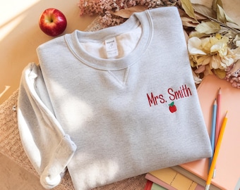 May include: A light gray sweatshirt with the embroidered text "Mrs. Smith" in red, along with a red apple graphic. The sweatshirt is folded on a soft, textured surface, with pencils and books nearby. A red apple and dried flowers are also in the image.