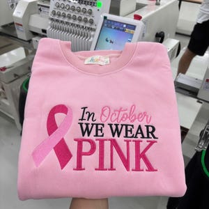 May include: A light pink crewneck sweatshirt featuring an embroidered dark pink breast cancer awareness ribbon on the left side. The text "In October WE WEAR PINK" is embroidered in a combination of cursive and block lettering across the front.