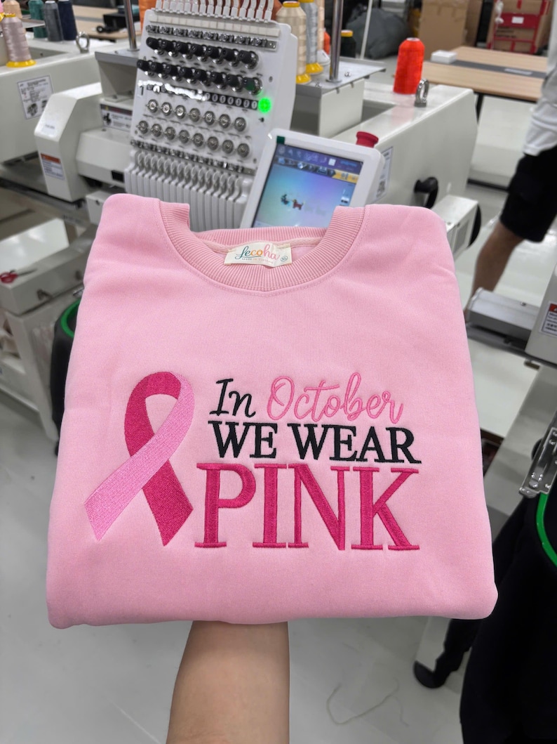 May include: A light pink crewneck sweatshirt featuring an embroidered dark pink breast cancer awareness ribbon on the left side. The text "In October WE WEAR PINK" is embroidered in a combination of cursive and block lettering across the front.
