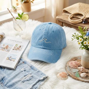 May include: A light blue baseball cap featuring a simple white embroidered wave design on the front panel. The cap has a structured crown, a curved brim, and eyelets for ventilation, crafted from a soft, washed-style fabric material.