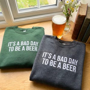 May include: Two sweatshirts, one forest green and one charcoal gray, each with the white text "IT'S A BAD DAY TO BE A BEER." A glass of beer sits nearby. The sweatshirts are on a wooden surface.