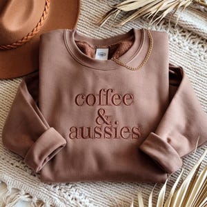 Könnte beinhalten: Ein braunes Sweatshirt mit dem gestickten Text "coffee & aussies". Das langärmlige Oberteil hat einen Rundhalsausschnitt und gerollte Manschetten. Eine goldene Kette ist über dem Ausschnitt drapiert. Ein brauner Hut ist im Hintergrund zu sehen.