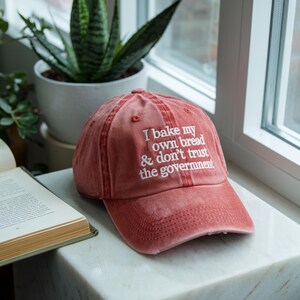May include: A red baseball cap with the embroidered text "I bake my own bread & don't trust the government" in white. The cap is made of a soft, worn material and is sitting on a marble surface.