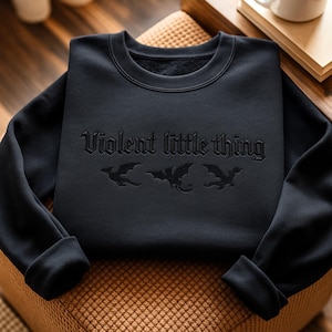 May include: Black sweatshirt with the phrase "Violent little thing" and three dragon silhouettes. The sweatshirt has a crew neck and long sleeves with cuffed wrists. The text and dragon graphics are in a darker shade of black.