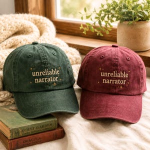 Puede incluir: Dos gorras de béisbol, una de color verde oscuro y otra granate, ambas con el texto "unreliable narrator" bordado en hilo dorado, acentuado con pequeños símbolos de estrellas encima y debajo de las palabras en el frente de cada gorra.