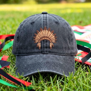 May include: A charcoal gray baseball cap with a distressed, washed-out finish. The front panel features an embroidered design of a brown feathered headdress with a geometric band pattern centered above the brim of the cap.