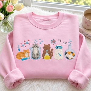 Peut inclure: Un sweat-shirt à col rond rose clair orné d'une broderie représentant cinq chats interagissant avec de la verrerie de laboratoire, des formules chimiques et des structures moléculaires. Le motif inclut un chat tigré, brun et tricolore.