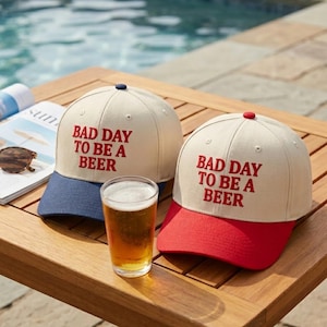 May include: Two baseball caps with the text "BAD DAY TO BE A BEER" in red. One cap has a blue peak, the other a red peak. A glass of beer sits in front of the caps on a wooden table, near a pool.