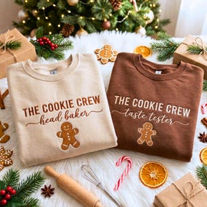 May include: Two crewneck sweatshirts, one beige and one brown, each featuring a gingerbread man graphic and embroidered text. The beige sweatshirt reads THE COOKIE CREW head baker, while the brown sweatshirt reads THE COOKIE CREW taste tester.