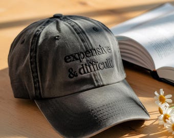 May include: A gray baseball cap with the embroidered text "expensive & difficult" in black. The cap is sitting on a wooden surface next to an open book and a few daisies.
