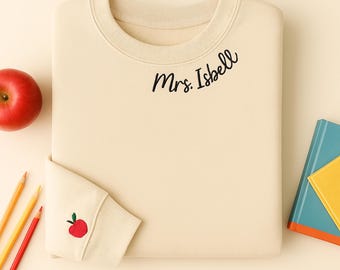 May include: Cream-colored sweatshirt with the name "Mrs. Isbell" in black script. The sleeve cuff features a small embroidered red apple. A red apple, pencils, and books are arranged around the sweatshirt.