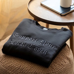 May include: A black sweatshirt with the embroidered text "Support Your Local Coven" in a gothic font. The sweatshirt is folded neatly on a woven pouf, with a small wooden side table and a notebook in the background.