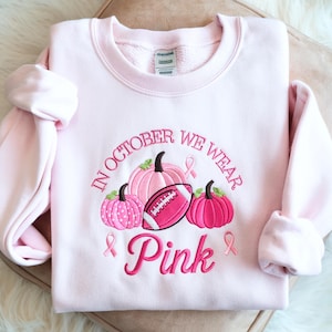 May include: A light pink crewneck sweatshirt featuring embroidered designs of three pumpkins, a football, and three breast cancer awareness ribbons. The text reads, "IN OCTOBER WE WEAR Pink" in dark pink embroidery. The garment has long sleeves.