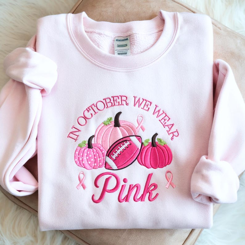 May include: A light pink crewneck sweatshirt featuring embroidered designs of three pumpkins, a football, and three breast cancer awareness ribbons. The text reads, "IN OCTOBER WE WEAR Pink" in dark pink embroidery. The garment has long sleeves.