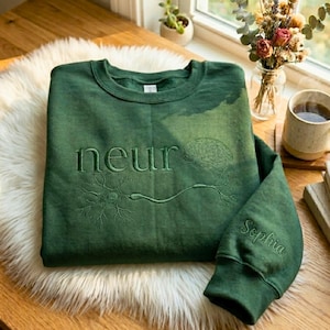 Könnte beinhalten: Ein dunkelgrünes Sweatshirt mit dem gestickten Wort "neur" und einem Gehirn- und Neuronen-Design. Auf dem Ärmel ist der Name "Sophia" in Grün gestickt. Das Sweatshirt liegt auf einem weißen Fellteppich.