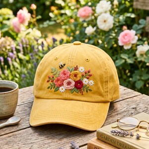 May include: A mustard yellow baseball cap featuring an embroidered floral arrangement on the front panel. The design includes pink, yellow, and red roses, small white daisies, green leaves, and a single small black bee hovering above the flowers.
