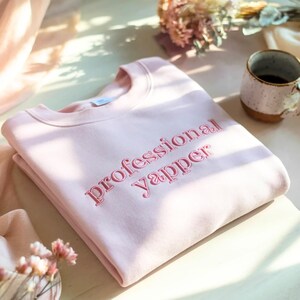 May include: A pale pink sweatshirt featuring the embroidered words "professional yapper" in pink thread. The garment is neatly folded. A coffee mug and flowers are visible in the background, creating a soft, inviting scene.