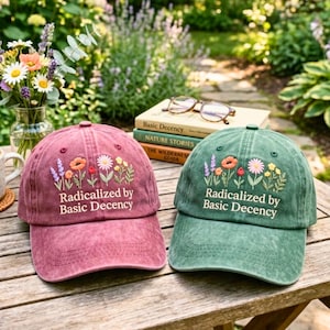 May include: Two baseball caps, one burgundy and one green, embroidered with colorful flowers and the text "Radicalized by Basic Decency." The caps are on a wooden surface, with books and glasses in the background.
