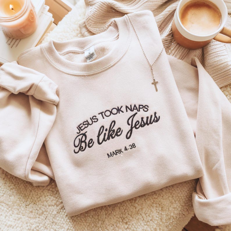 May include: A pale pink sweatshirt with the embroidered text "JESUS TOOK NAPS Be like Jesus MARK 4:38". A gold cross pendant hangs on the front. A lit candle and a coffee cup are visible in the background.