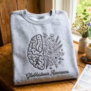 Puede incluir: Una sudadera gris con una ilustración en blanco y negro de un cerebro y un diseño floral. Las palabras "Glioblastoma Awareness" están impresas debajo del gráfico. La sudadera está doblada sobre una superficie de madera.