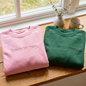 May include: Two crewneck sweatshirts, one light pink and one dark green, each featuring the embroidered text "born to yap." in a matching thread colour. The garments have long sleeves, ribbed necklines, and a soft, fleece-like texture throughout.