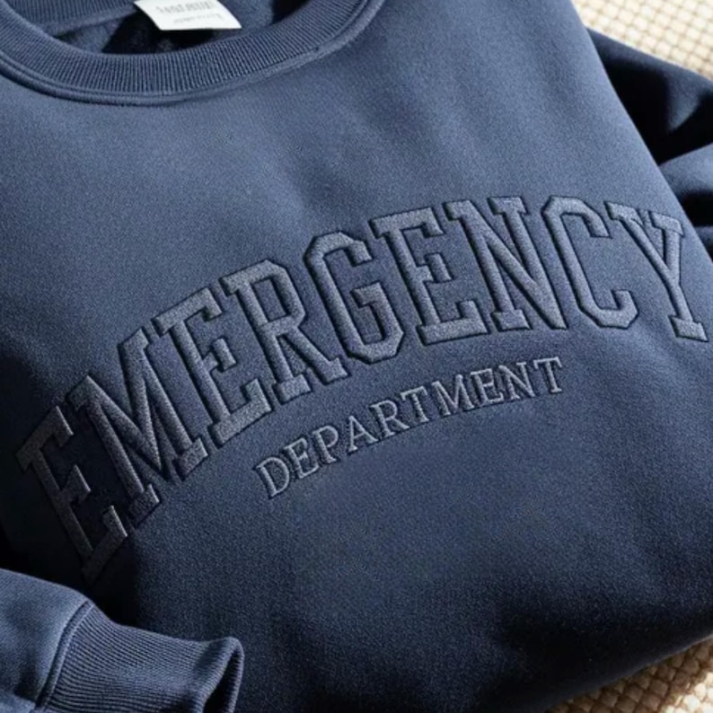 K&ouml;nnte beinhalten: Marineblaues Sweatshirt mit der gestickten Aufschrift "EMERGENCY DEPARTMENT" in passendem dunklerem Blau. Das Sweatshirt hat einen Rundhalsausschnitt und wirkt weich und bequem.