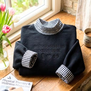 May include: A black long-sleeve sweatshirt featuring a mock neck collar and sleeve cuffs in a black and white gingham pattern. The front is embroidered with the text MOVEMENT IS MEDICINE, accompanied by icons of a spine, a heart, and a dumbbell.