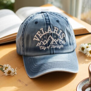 May include: A blue denim baseball cap with the word "VELARIS" embroidered in white, along with a mountain graphic and the text "City of Starlight". The cap is sitting on a wooden surface.