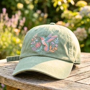 Puede incluir: Una gorra de béisbol color verde salvia con un diseño bordado de un colibrí en vuelo rodeado de pequeñas flores y hojas coloridas. El bordado incluye tonos de rosa, naranja, azul y morado en la parte frontal de la gorra.