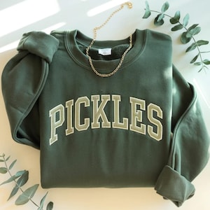 以下が含まれることがあります： 「PICKLES」の文字がライトグリーンで刺繍された、ダークグリーンのスウェットシャツ。 金色のチェーンネックレスがスウェットシャツの上に置かれています。 袖口は折り返されており、背景には緑の小枝があります。