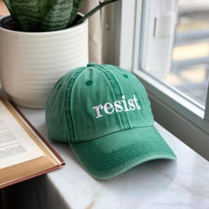 Puede incluir: Gorra de béisbol verde con la palabra "resist" bordada en blanco. La gorra está hecha de una tela suave y tiene una visera curva. El sombrero está sobre una superficie de mármol.