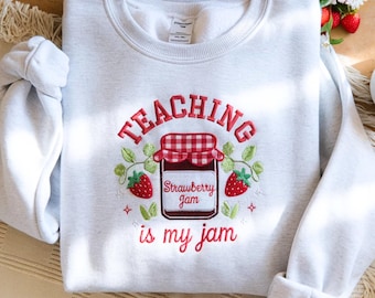 May include: A light gray sweatshirt with the embroidered phrase "TEACHING is my jam." The design features a strawberry jam jar with a red and white checkered lid, surrounded by strawberries and green leaves.