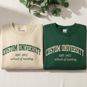 May include: Two crewneck sweatshirts, one beige and one dark green. Both feature the embroidered text "CUSTOM UNIVERSITY EST. 1972 school of nursing" in a contrasting color. The sweatshirts are displayed on a neutral surface with leafy green sprigs.