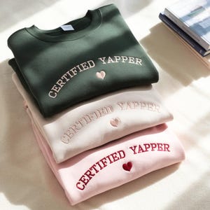 May include: Three sweatshirts in green, beige, and pink are stacked. Each features the embroidered text "CERTIFIED YAPPER" and a heart. The sweatshirts are neatly folded and arranged in a pile, showcasing the embroidered design.