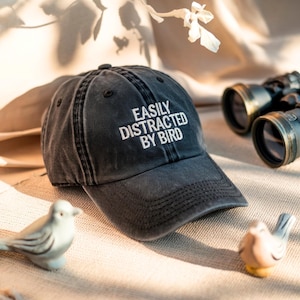 May include: A black, distressed baseball cap with the text "EASILY DISTRACTED BY BIRD" in white. The cap is on a beige surface, with a pair of binoculars and two small bird figurines nearby. The cap has a curved brim.