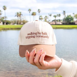 May include: A beige and brown baseball cap with the text "walking by faith tripping constantly" embroidered in brown. The cap is held up in front of a lake and palm trees.