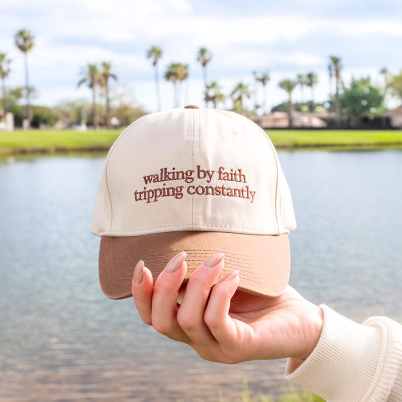 May include: A beige and brown baseball cap with the text "walking by faith tripping constantly" embroidered in brown. The cap is held up in front of a lake and palm trees.