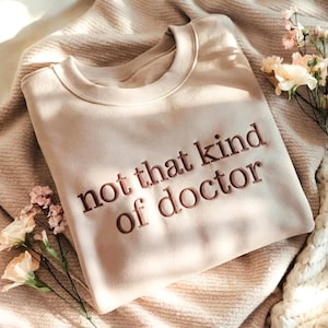 May include: A beige sweatshirt with the embroidered text "not that kind of doctor" in a darker brown color. The sweatshirt is folded on a textured blanket, with flowers in the background.