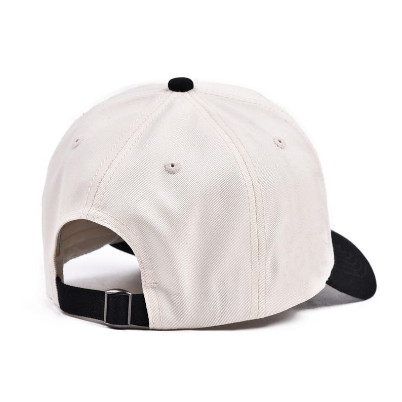 May include: A cream-colored baseball cap with a black brim and button. The adjustable strap is black with a silver-colored metal buckle. The hat is made of a sturdy fabric.