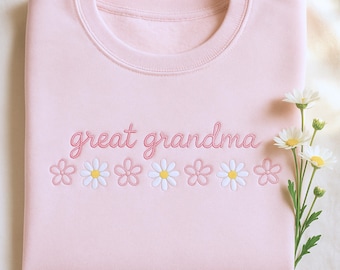 May include: A light pink sweatshirt with the embroidered text "great grandma" in a cursive font. Below the text are embroidered daisies with white petals and yellow centers. The sweatshirt is folded.