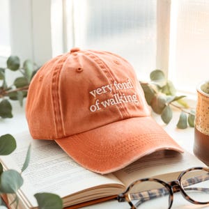 May include: An orange baseball cap with the embroidered text "very fond of walking" in white. The hat is sitting on an open book with eyeglasses in front of a window.