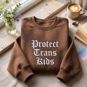 May include: A brown sweatshirt with the words "Protect Trans Kids" in white gothic lettering. The sweatshirt is folded on a wooden surface, next to a cup of coffee with latte art, a small vase of flowers, and a book.