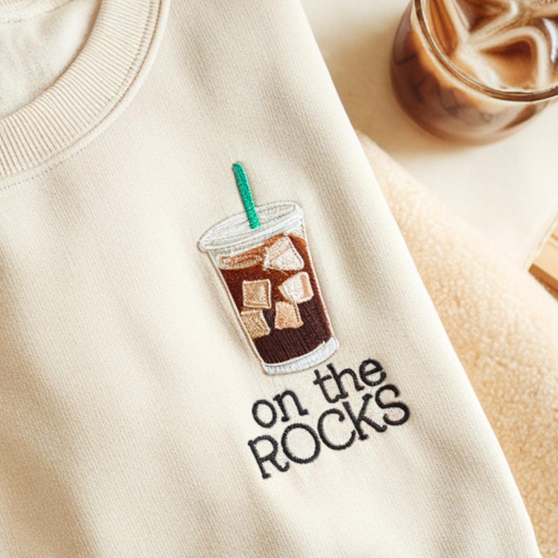 May include: Cream-colored sweatshirt with an embroidered iced coffee design. The design features a clear cup with brown coffee, ice cubes, and a green straw. The words "on the ROCKS" are embroidered below the coffee cup.