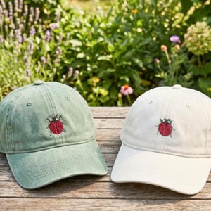 May include: Two baseball caps, one in sage green and one in off-white, each featuring a small, embroidered red ladybug with black spots and details centered on the front panel. Both caps have a curved brim and a classic six-panel construction.