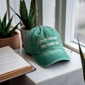 May include: A green baseball cap with the embroidered text "have the day you voted for." The hat is sitting on a white marble surface next to a book and potted plants. The hat has a curved brim and a classic six-panel design.